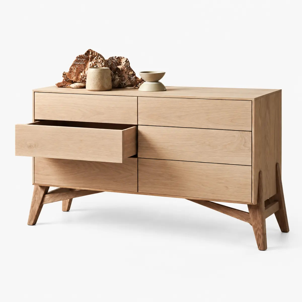 A mid-century modern wooden dresser with a natural oak finish, featuring six drawers (one open on the left), angled legs, and decorative ceramic objects on top, set against a plain white background.