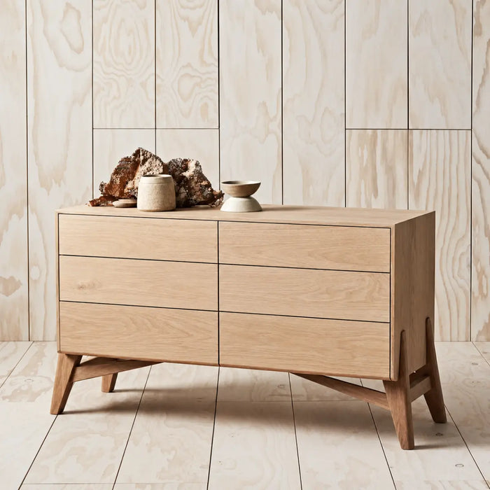 A mid-century modern wooden sideboard with six drawers, styled with decorative objects on top, set against a light wood paneled wall and floor.